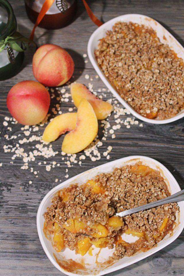 A topdown photo of two individual peach crumbles in white oval ramekins with a couple of whole peachs to the left of the dishes. 