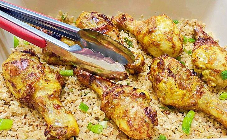 A horizontal photo of the chicken legs laying on cauliflower rice with a red pair of tongs.