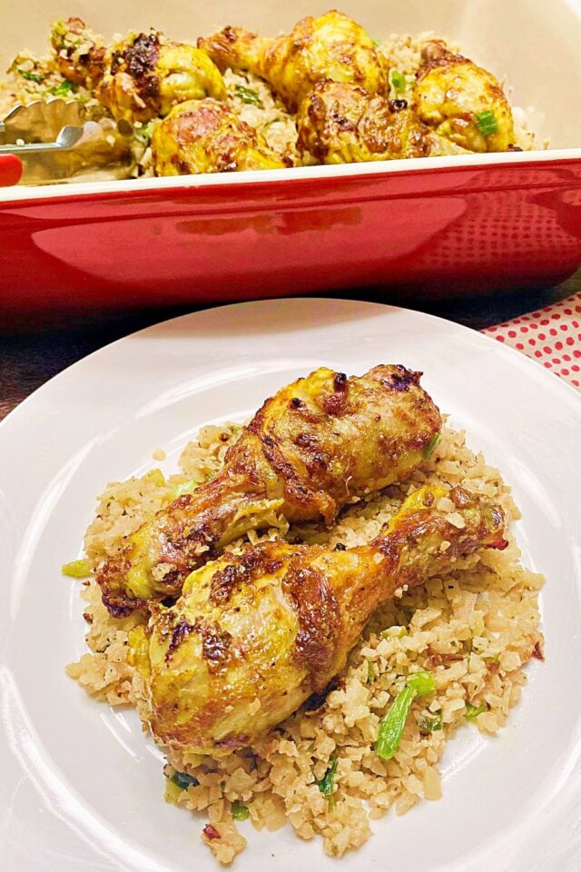 Two chicken legs on a be of cauliflower rice served on a white plate with a red casserole dish in the background. 