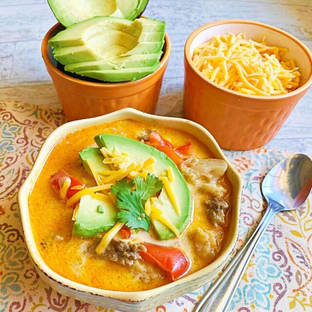 A bowl of fajita soup with two smaller bowls beside it full of cheese and avocado for toppings. 