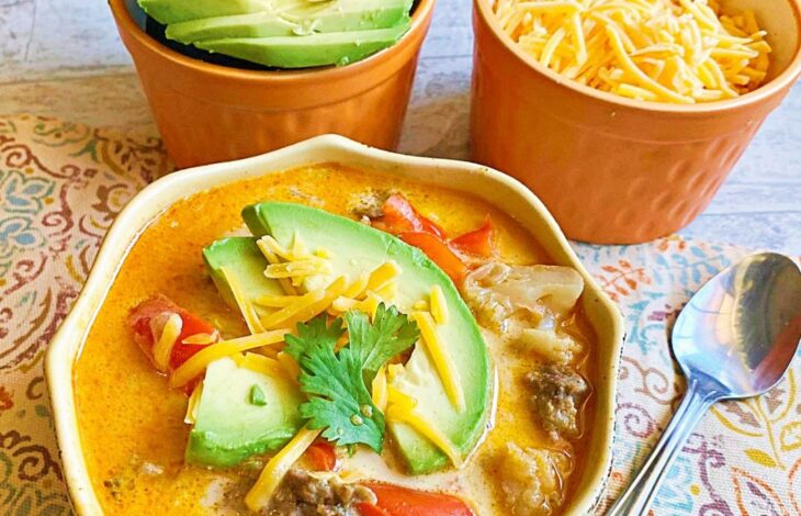 A bowl of fajita soup with two smaller bowls beside it full of cheese and avocado for toppings.