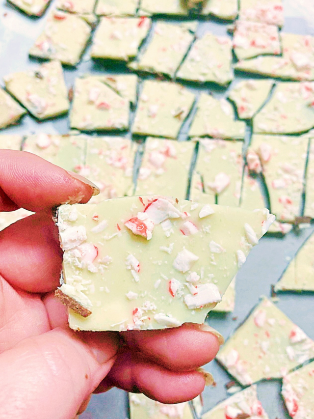 A hand holding a piece of peppermint bark with more pieces in the background on parchment paper.
