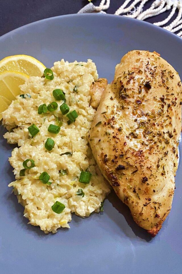 A whole grilled chicken breast with some cauliflower rice and lemon wedges on a gray plate