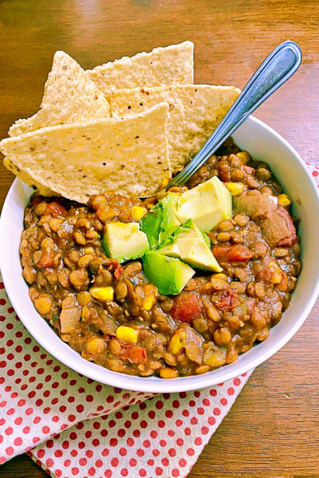 A white round bowl of lentil chili with a spoon and a handful of tortilla chips. 