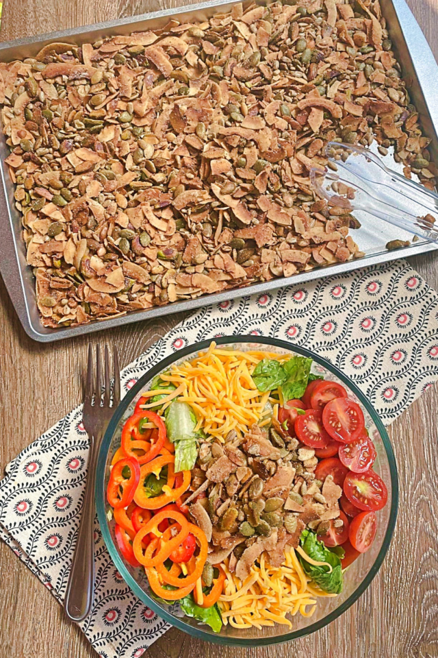 A top down view of a pan of the salad toppers mix on a baking sheet with a bowl of salad sitting to the front of it.