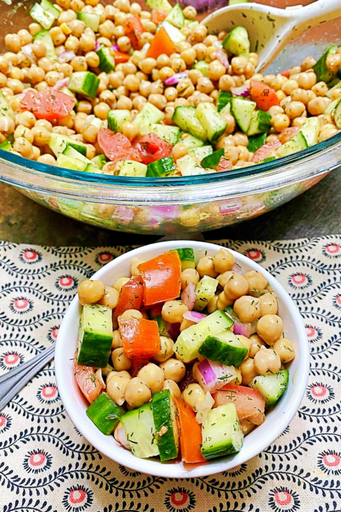 A large glass bowl in the back ground full of chickpea salad with a small white bowl sitting in front of it loaded with a serving of salad. 