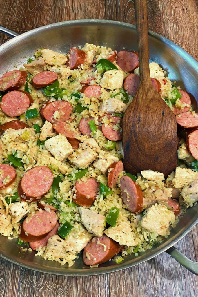 A top down photo of a silver skillet full of Chicken and Sausage Skille recipe and a brown wooden serving spoon in the top right of the photo.