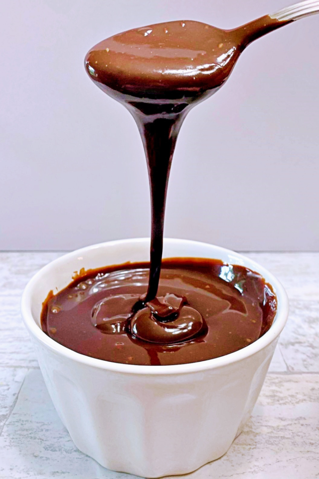 A small white bowl of sugar free chocolate fudge sauce with a spoon full drizzling done into the bowl.