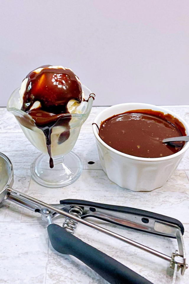A photo of vanilla ice cream in a clear sundae cup topped with the sugar free chocolate fudge sauce. An ice cream scoop and bowl of chocolate are also in the photo.