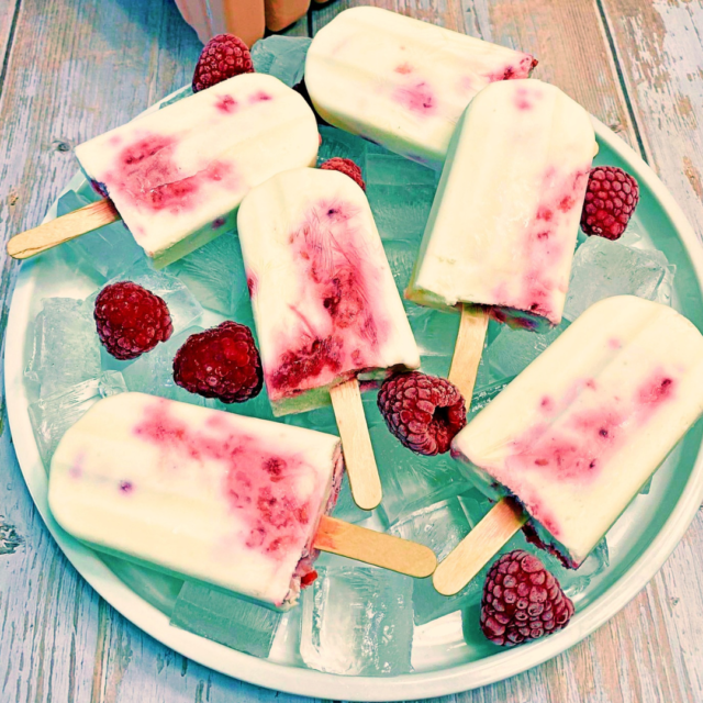 An icy plate of Raspberry Cheesecake Popsicles surrounded by fresh red raspberries.