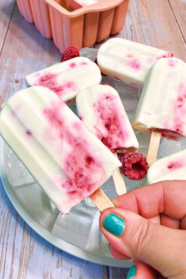 A hand holding a Keto Raspberry Cheesecake Popsicle with a full plate of popsicles in the background. 