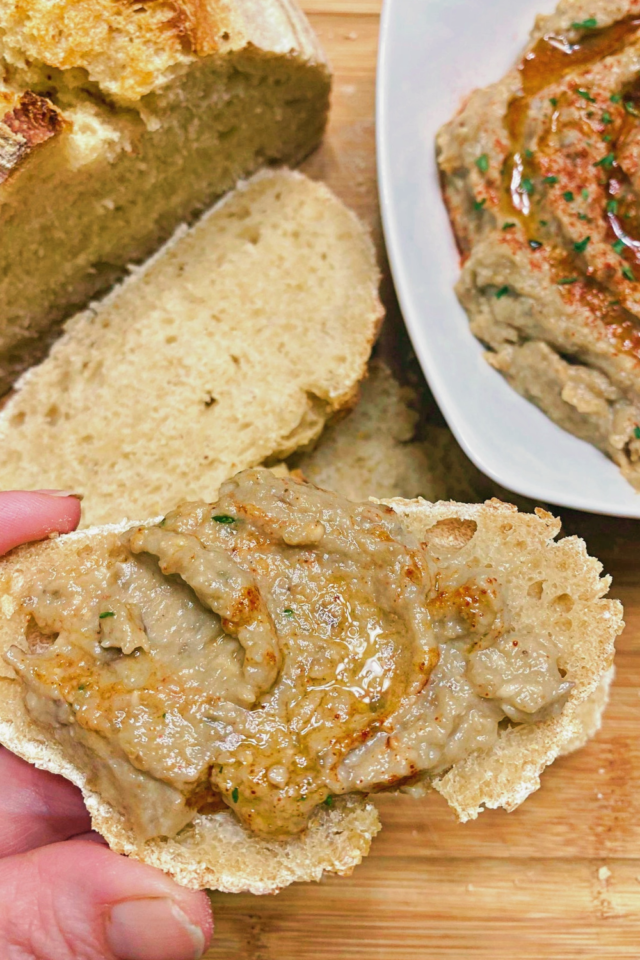 A piece of bread spread with eggplant dip in the foreground and more bread and dip in the background. 