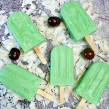 Five tropical limeade popsicles laying on top of coconut flakes with cherries laying around.